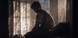 A seated figure silhouetted against a curtained window.